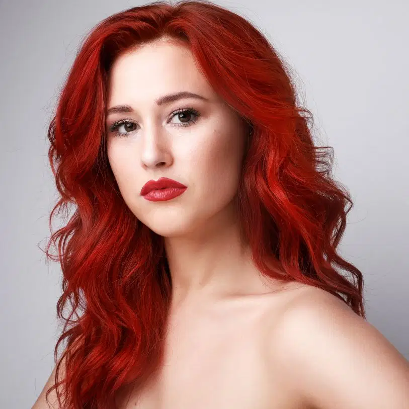 Mulher de pele clara e longos cabelos ondulados tom vermelho-vibrante usa batom vermelho marcante e olha fixamente para a câmera