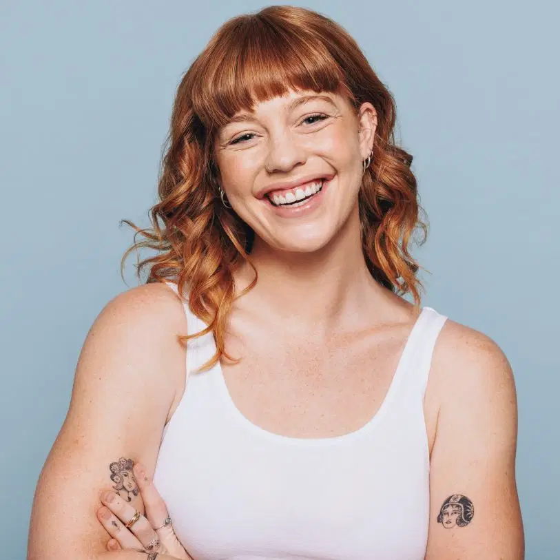 Mulher ruiva sorridente de franja e cabelo ondulado médio, vestindo regata branca, posa de braços cruzados contra um fundo azul-claro.