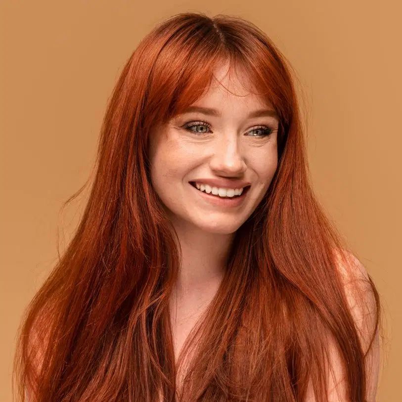 Mulher ruiva de cabelos longos e lisos com franja sorri abertamente, posando contra um fundo bege-terroso
