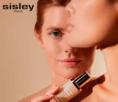 Mulher jovem com olhos verdes intensos e pele brilhante segurando um frasco de base Sisley.