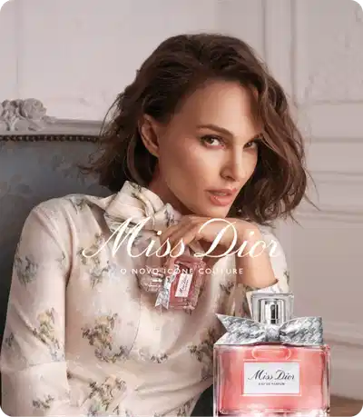 Mulher sentada em poltrona cinza, usando blusa estampada em tons neutros, segurando frasco de Miss Dior Eau de Parfum. Ao lado, frasco rosado Miss Dior. Texto em branco: Miss Dior, o novo ícone couture.