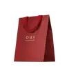 Sacola de presente vermelha em material texturizado, com alças e detalhes na mesma cor. Apresenta o logotipo "O.U.i" em letras douradas. A embalagem tem um design minimalista e elegante, com o vermelho vibrante como cor predominante.