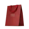 Sacola de presente vermelha em material texturizado, com alças e detalhes na mesma cor. Apresenta o logotipo "O.U.i" em letras douradas. A embalagem tem um design minimalista e elegante, com o vermelho vibrante como cor predominante.