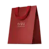 Sacola de presente vermelha em material texturizado, com alças e detalhes na mesma cor. Apresenta o logotipo "O.U.i" em letras douradas. A embalagem tem um design minimalista e elegante, com o vermelho vibrante como cor predominante.