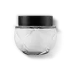 Frasco de vidro transparente arredondado com tampa preta. A embalagem tem um design minimalista e clean, com linhas suaves e uma forma esférica. As cores predominantes são o preto da tampa e o transparente do vidro e a cor branca do creme.