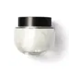 Frasco de vidro arredondado, transparente com tampa preta de rosquear, contendo o creme na cor branca. Design minimalista e elegante.