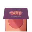 A imagem mostra o produto Blush Me da marca Océane. É uma embalagem quadrada de cor roxa que está semiaberta mostrando a cor do blush em um tom laranja com leve brilho perolado. A parte interna da embalagem é de cor pêssego claro,