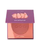 A imagem mostra o produto Blush Me da marca Océane. É uma embalagem quadrada de cor roxa que está semiaberta mostrando a cor do blush em um tom rosa com leve brilho perolado. A parte interna da embalagem é de cor pêssego claro,
