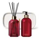 Três produtos da marca Casa 214 Gardênia Imperial: dois vidros vermelhos, sendo um difusor de ambientes com valetas e o outro com bico dosador preto de sabonete líquido e uma badeja de lavabo branca com logo da marca O Boticário em dourado.