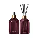 A imagem mostra dois vidros retangulares roxos, com rótulo em dourado. Um deles é o aromatizador de ambientes com válvula spray preta e o outro é um difusor com tampa dourada que acompanha varetas de madeira. Ambos são a linha Casa 214 Figo Magnífico.