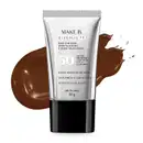 A imagem mostra um tubo de base líquida Make B. Glycolic TX. Ele é prateado, possui tampa preta e informações em letras pretas. Atrás da embalagem está a textura do produto, mostrando sua intensidade e a real cor da base.