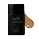 Frasco preto de base da linha Make B. Mate Salicylic com texto em branco trazendo informações do produto, como presença de ácido salicílico, controle de oleosidade e resistência ao suor. Atrás da embalagem, vemos um pouco de base bege clara espalhada.