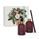 Caixa branca com ilustração floral e dois vidros retangulares na cor roxa, com rótulo em dourado. Um deles é o aromatizador de ambientes com válvula spray preta e o outro é um difusor com tampa dourada que acompanha varetas de madeira pretas.