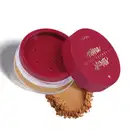 A imagem mostra a embalagem de um pó facial solto, redonda de plástico transparente com parte superior vermelha de furinhos e tampa vermelha com logo da marca escrito em dourado na lateral. No fundo, próximo ao produto, uma amostra da cor e textura do pó.