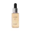 Primer líquido. Frasco transparente com tampa prateada e preta contendo um líquido dourado brilhante com partículas cintilantes. Rótulo dourado com o nome do produto e da linha. Embalagem simples destacando o aspecto metálico e iluminador do produto.