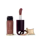 O produto de beleza Eudora Make é um batom líquido com acabamento cremoso na cor marrom avermelhado, em embalagem com frasco e aplicador. Possui rótulo com o nome "Kiss Me Tint Charm" e detalhes decorativos em dourado.