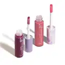 Imagem de dois frascos de gloss labial, um roxo com título Cachos de Uva e outro rosa com título Doces Delírios. Ambos em recipiente transparente com aplicador em forma de esponja na mesma cor do gloss. O fundo da imagem é branco com sombreado.