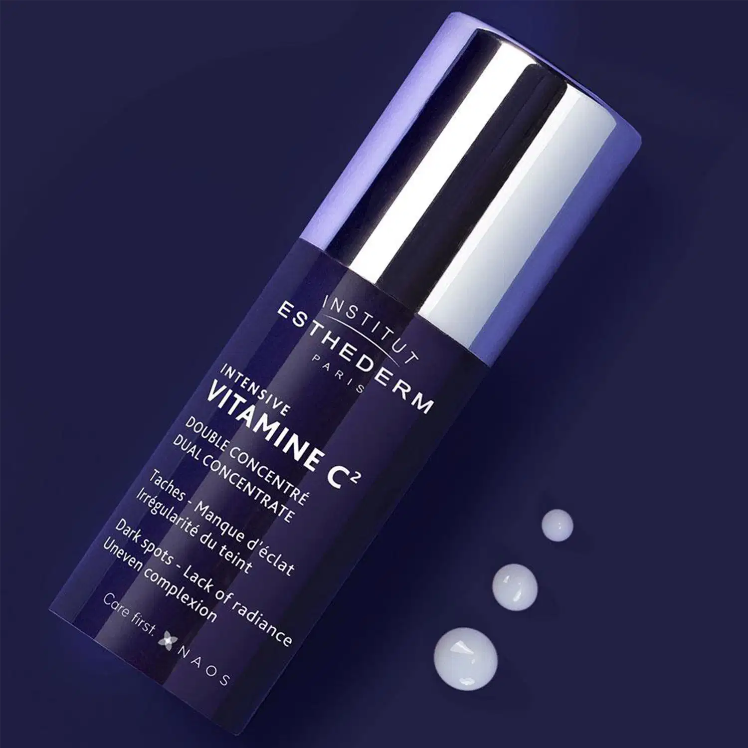 Embalagem de produto de beleza da marca Esthederm, com frasco de vidro escuro e tampa prateada, em fundo azul escuro com pequenas esferas brancas, sugerindo um ambiente sofisticado e de cuidados com a pele.