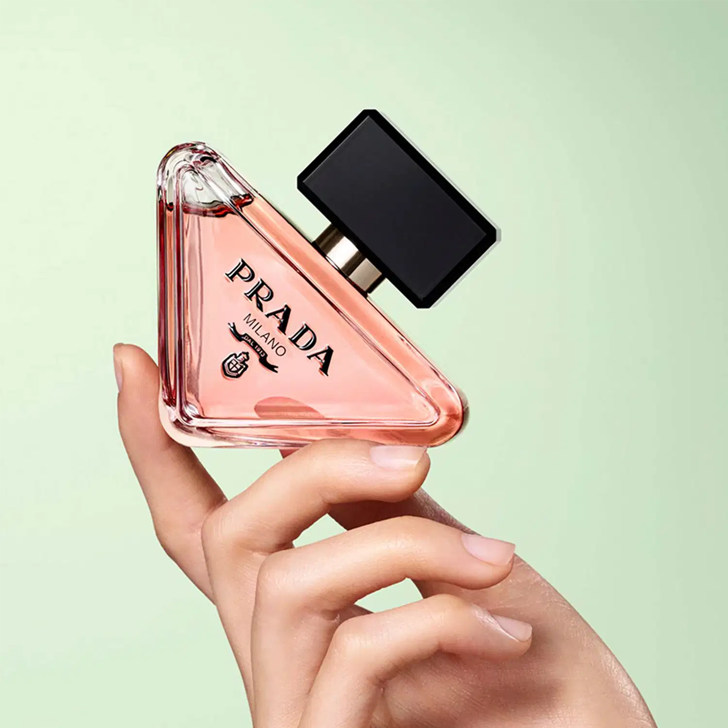 Frasco de perfume Prada com design triangular em vidro transparente, contendo líquido de cor rosa clara. O rótulo preto destaca o nome da marca "PRADA" em letras brancas.