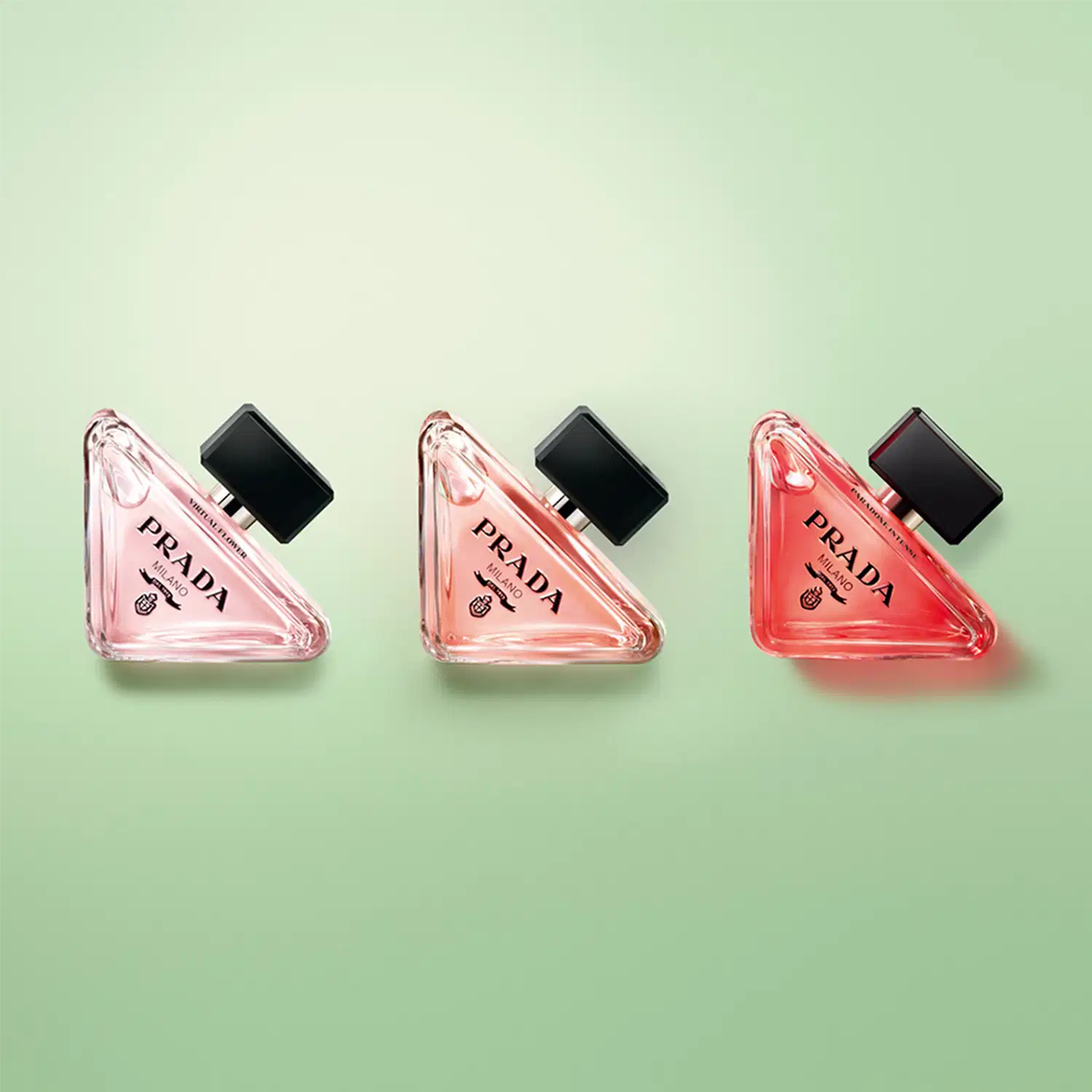 Três frascos de perfume Prada em diferentes tons de rosa, com tampas pretas e o logotipo da marca visível. Os frascos possuem formas triangulares e elegantes, transmitindo uma imagem sofisticada e luxuosa.