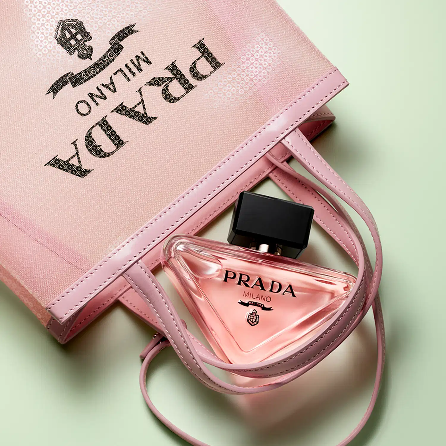Frasco de perfume Prada com design triangular em vidro transparente, contendo líquido de cor rosa clara. O rótulo preto destaca o nome da marca "PRADA" em letras brancas.
