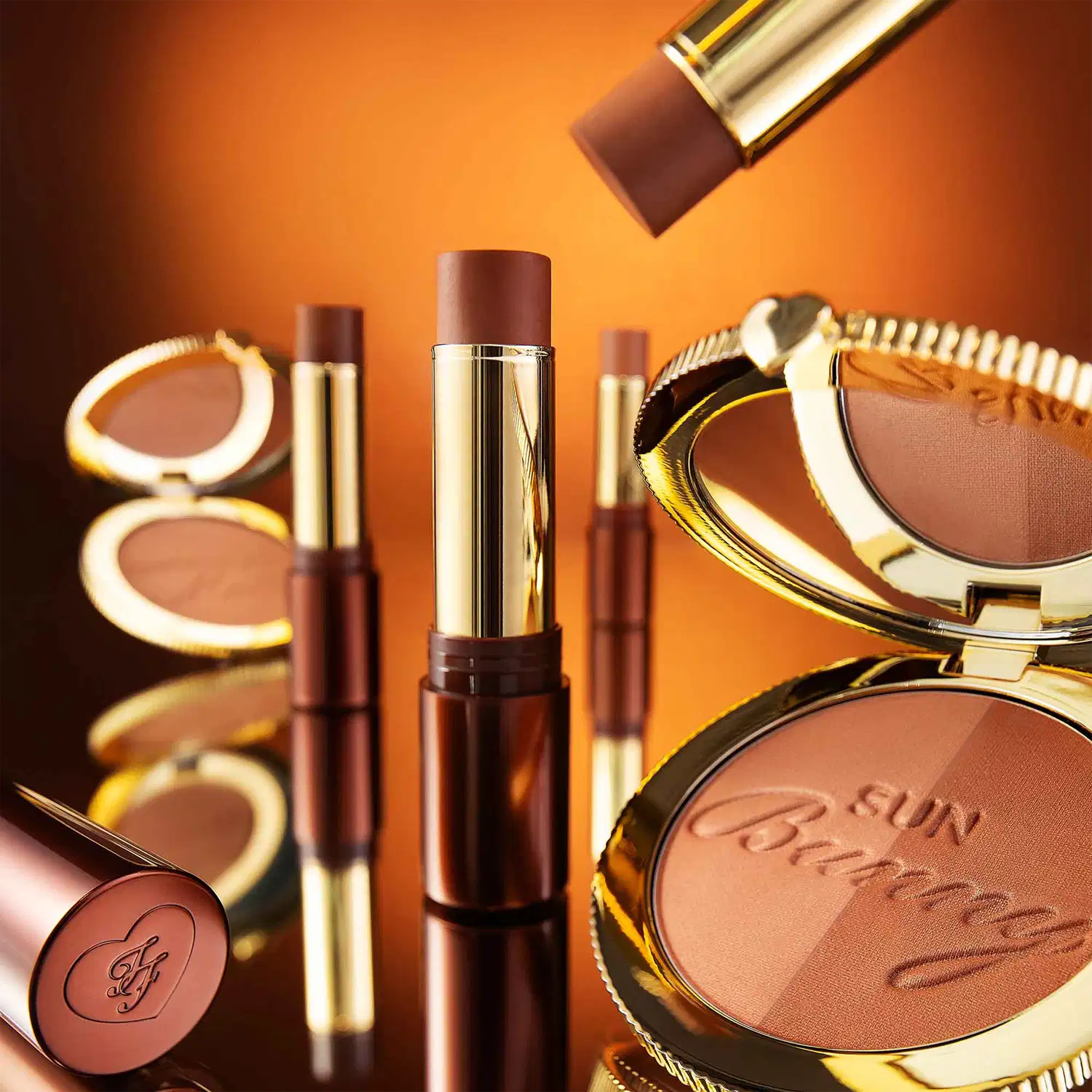 Conjunto de produtos de maquiagem da marca Too Faced. Inclui bronzers em embalagens douradas e marrons, pó compacto bronzeador com a inscrição Sun Bunny, pó compacto de blush com o logotipo Too Faced, sombras compactas em tons terrosos.