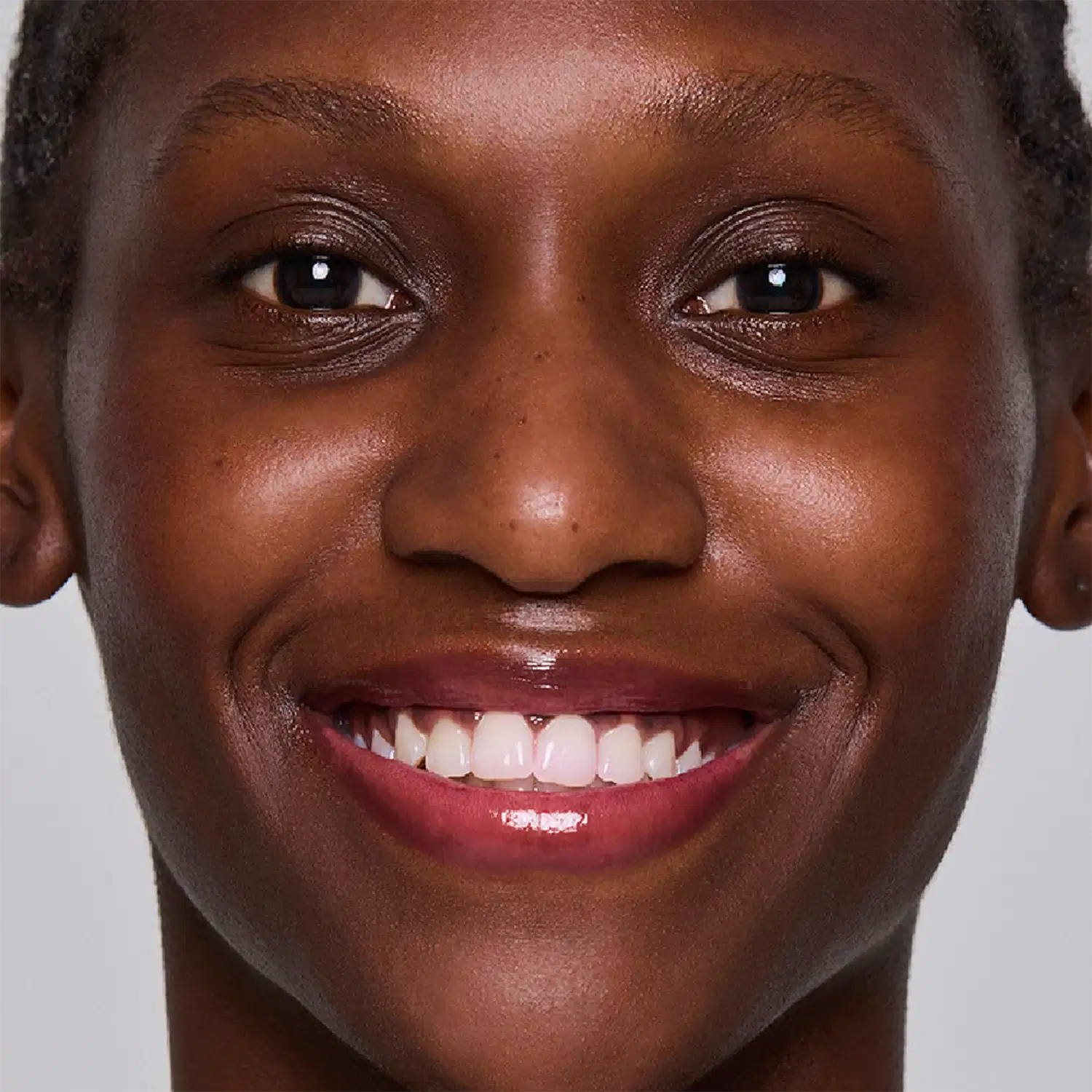 A imagem apresenta uma modelo de pele negra com a pele ao natural. Apresenta um lindo sorriso com dentes brancos e os lábios bem hidratados, levemente rosados e com brilho suave.