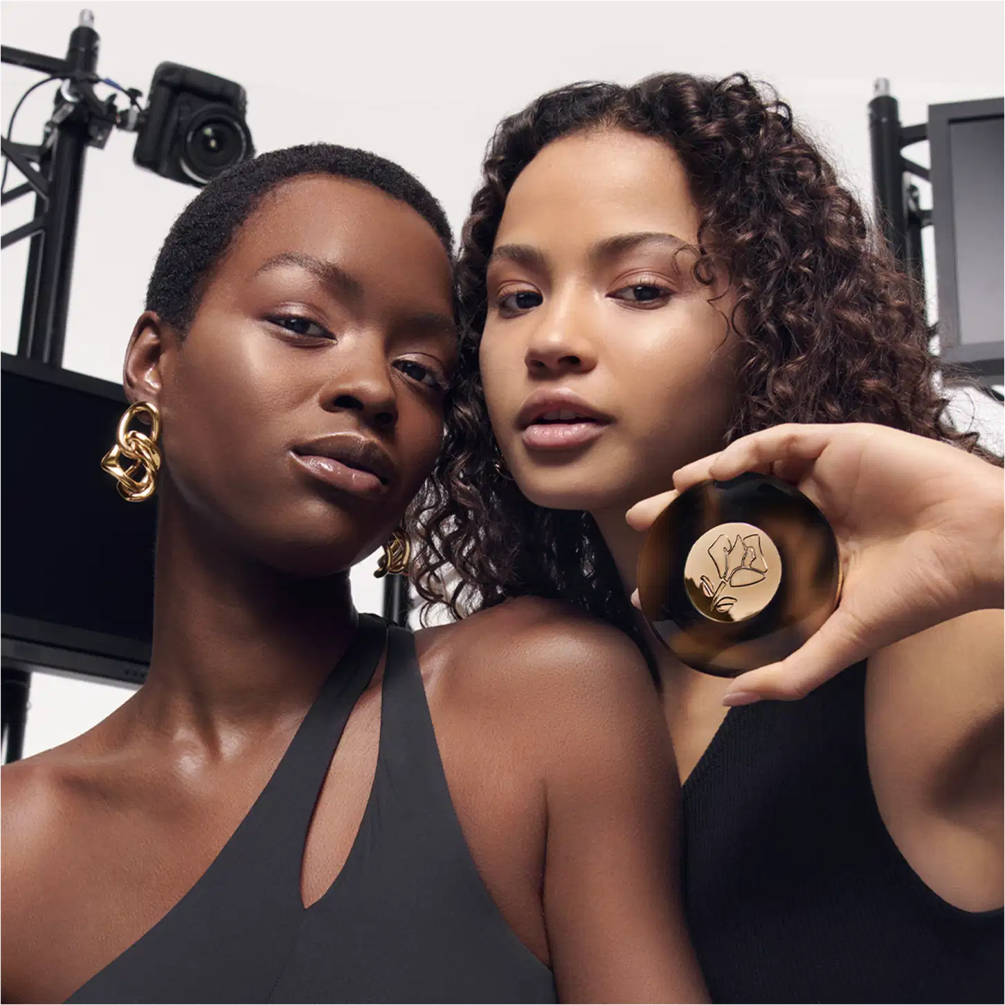 A imagem traz uma modelo de cabelo cacheado e outra de cabelo curtinho, posando em um cenário com equipamentos fotográficos ao fundo. Uma delas carregam o iluminador na mão mostrando a embalagem e a outra posa para a foto com uma expressão serena.