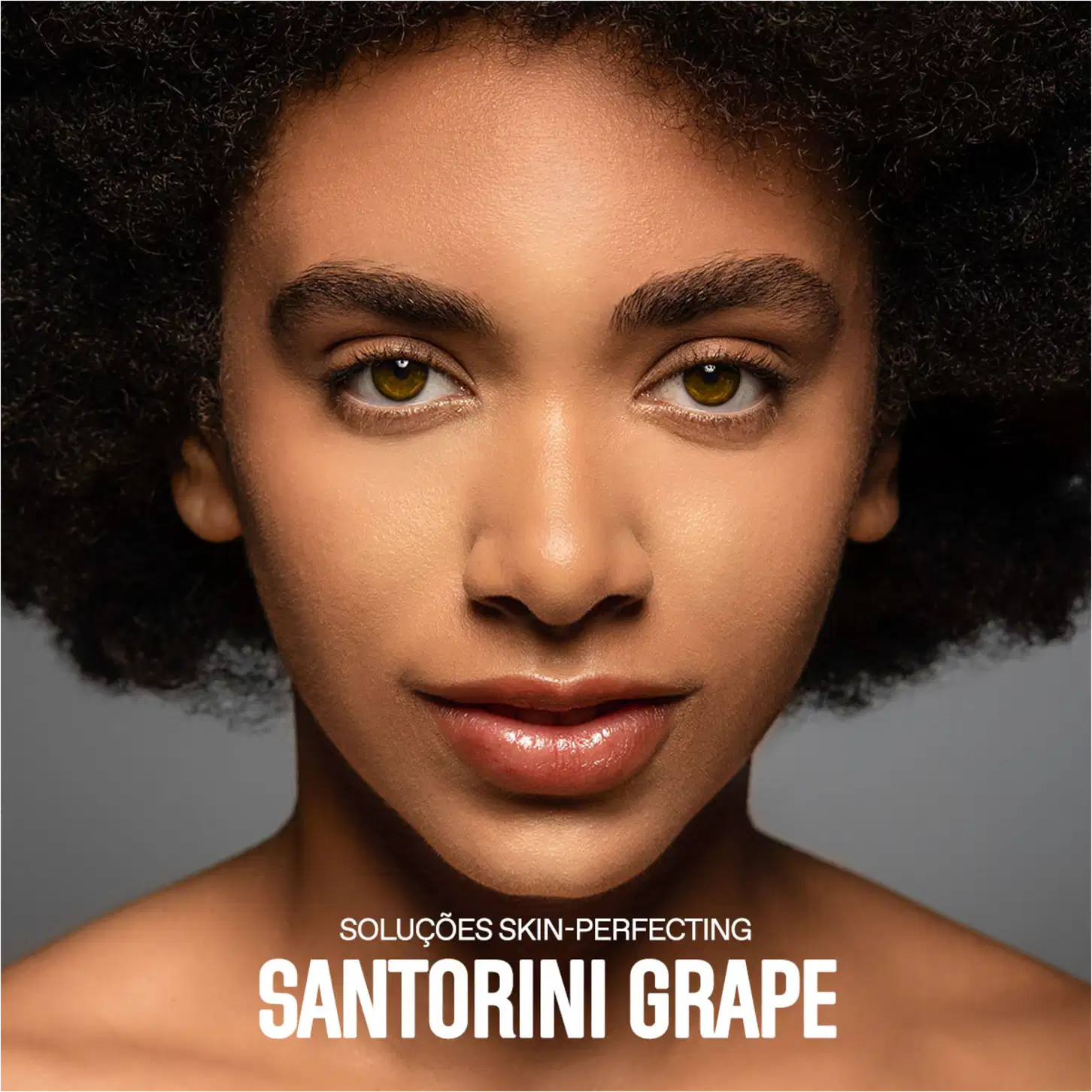 A imagem mostra um close-up do rosto de uma mulher negra com cabelos crespos e curtos. Ela tem olhos amendoados de cor amarelo-esverdeada. Sua pele tem um aspecto aveludado e uniforme, provavelmente resultado do uso dos produtos anunciados.