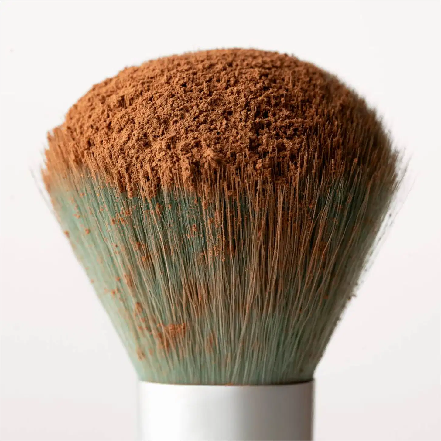 Pincel de maquiagem com cerdas densas e macias de cor marrom avermelhada. O cabo é branco e a base é verde musgo com detalhes em tom ferrugem. O formato é arredondado e denso, indicando ser um pincel para aplicação de pó ou blush.