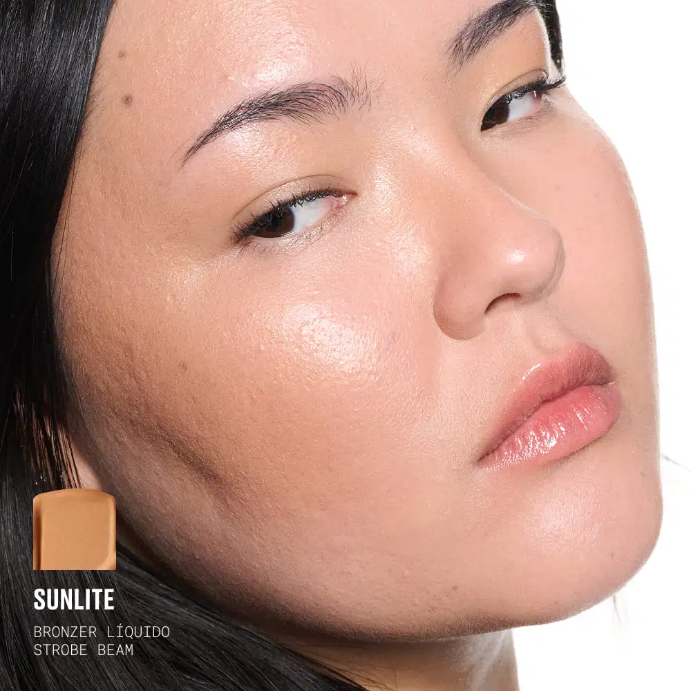 A imagem mostra uma mulher asiática com cabelos escuros usando a cor Sunlite do bronzer líquido Strobe Beam da MAC, destacando o efeito iluminador.