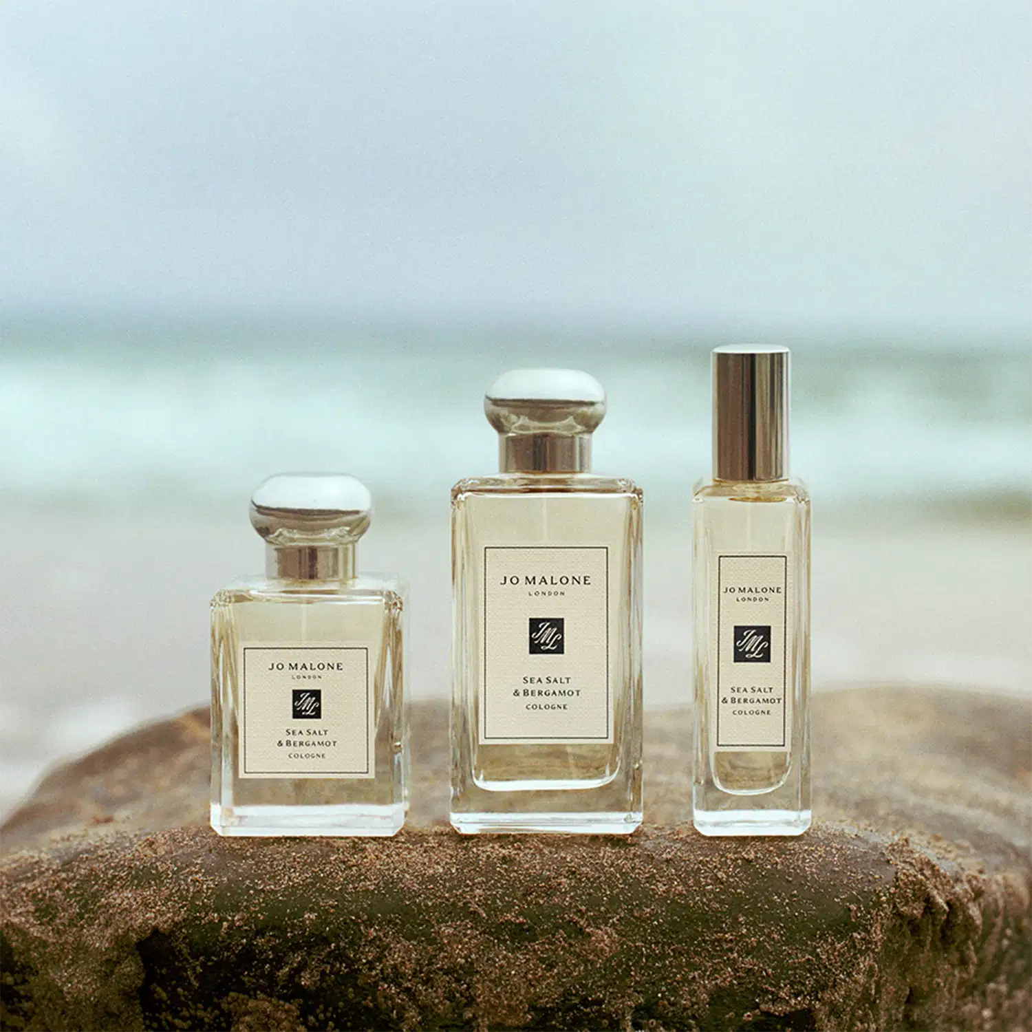 Três frascos de perfume da marca Jo Malone London, com a fragrância Sea Salt & Bergamot, em diferentes volumetrias, dispostos sobre uma rocha em um ambiente natural, com o mar ao fundo, sugerindo um cenário costeiro e relaxante.