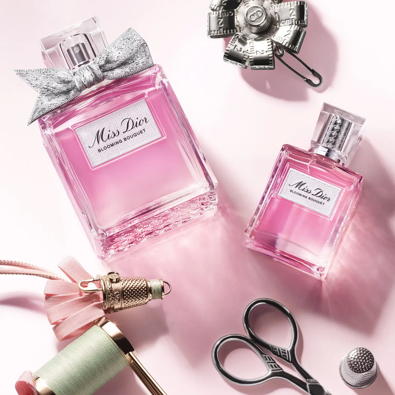 Dois frascos de Miss Dior Blooming Bouquet em vidro rosa sobre superfície rosa claro, acompanhados de acessórios decorativos em metal prateado e rosa, incluindo tesoura, carretel de linha e aplicador de perfume.