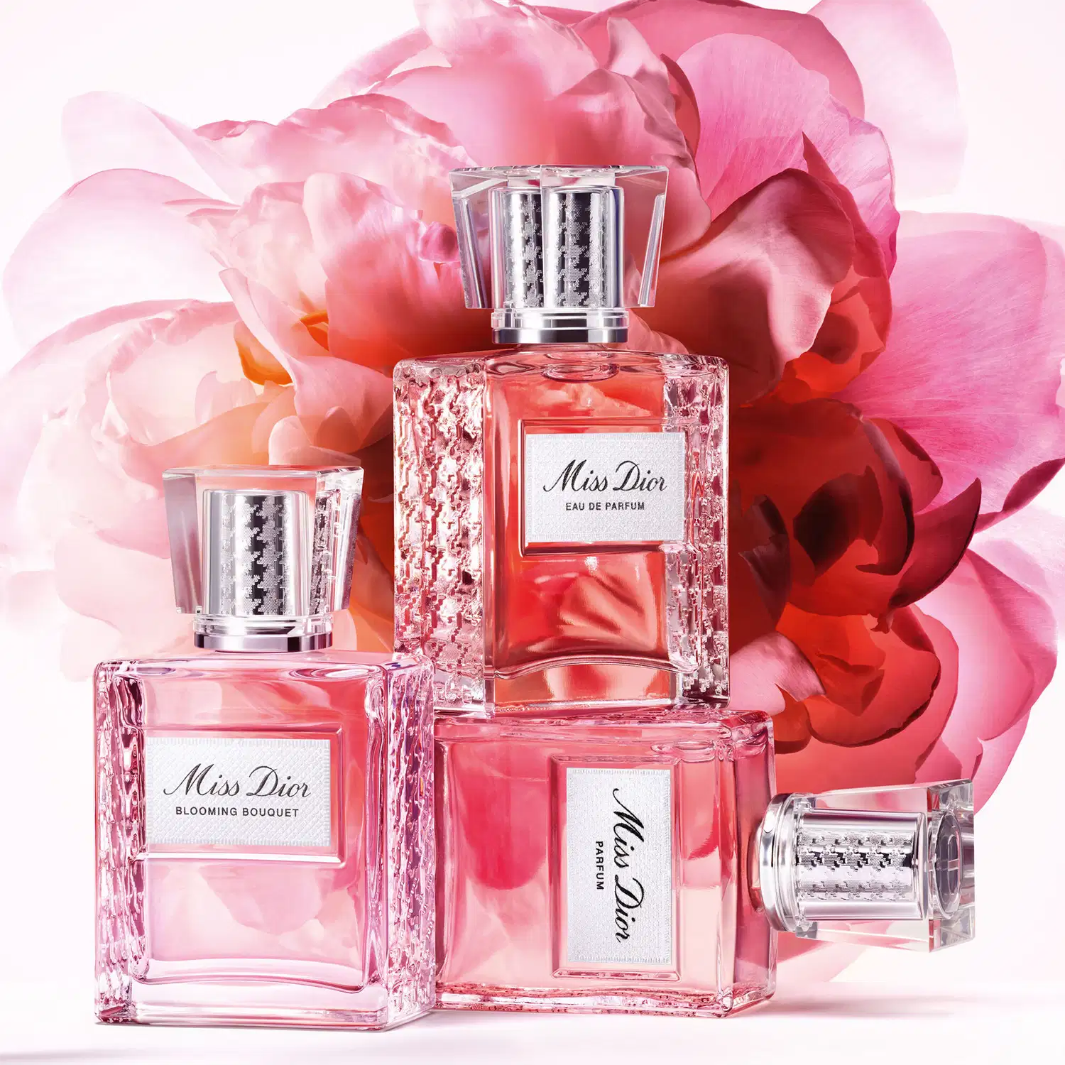 Três frascos de item de perfumaria Dior em vidro cristalino com rótulos brancos, dispostos sobre pétalas de rosa rosa e vermelha, com flores ao fundo desfocadas.