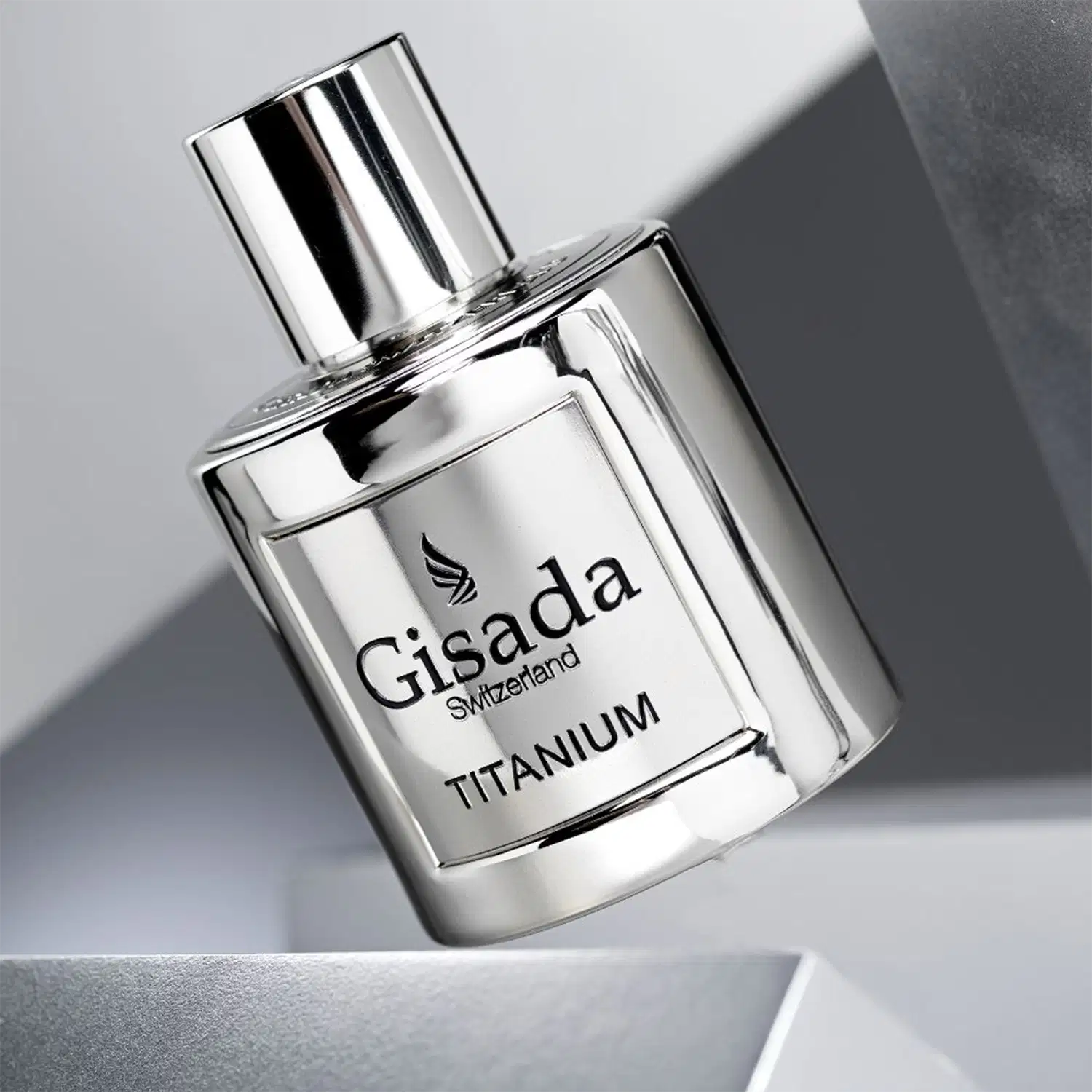 Perfume Gisada Titanium em frasco de vidro transparente com tampa prateada cilíndrica. Embalagem branca e prata com logo alado da marca e texto "Gisada Switzerland TITANIUM" em preto. Design minimalista e elegante.