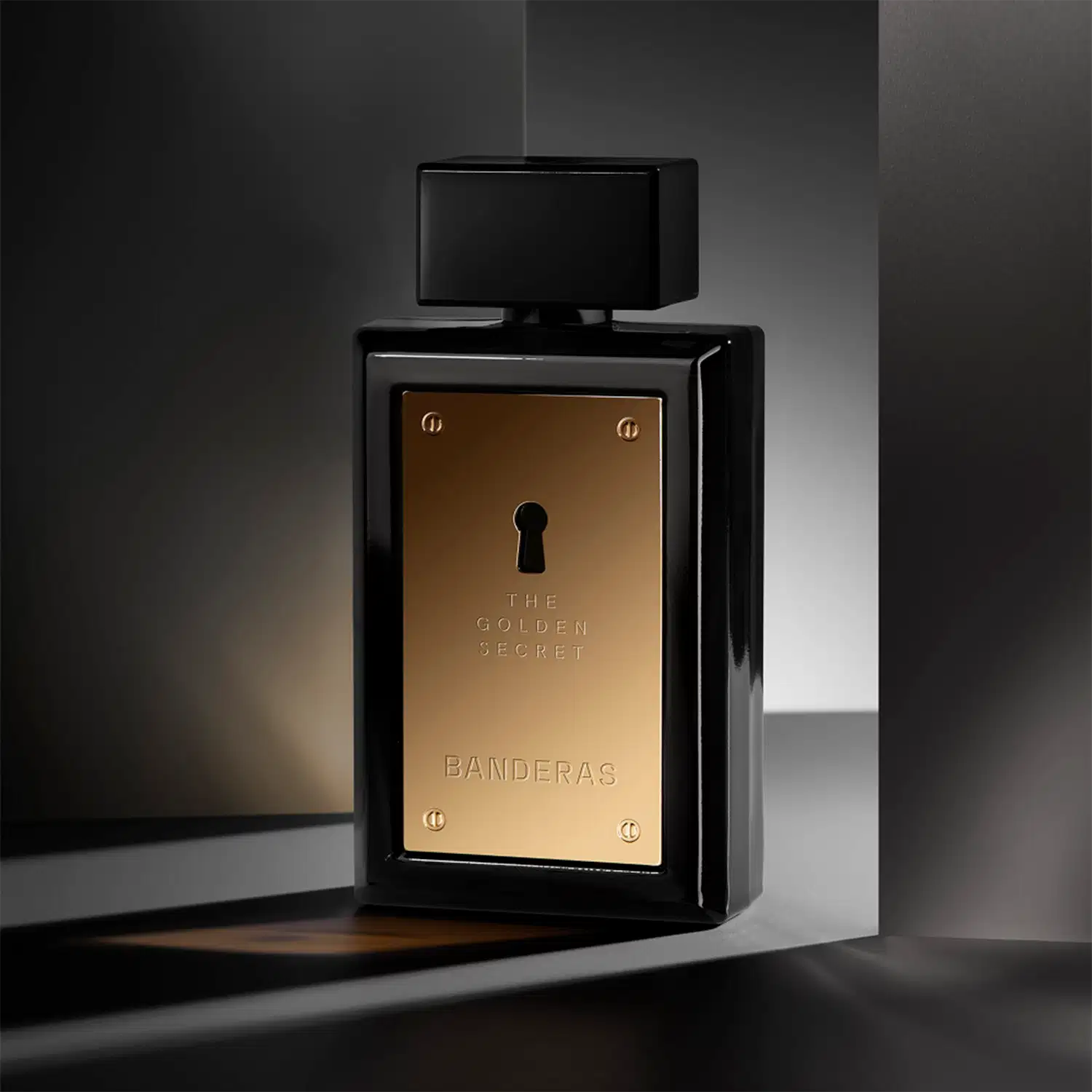 Frasco de perfume preto com painel frontal dourado exibindo o texto "The Golden Secret" e "Banderas" em letras douradas. O frasco possui um design elegante e minimalista, com um pequeno orifício no centro.