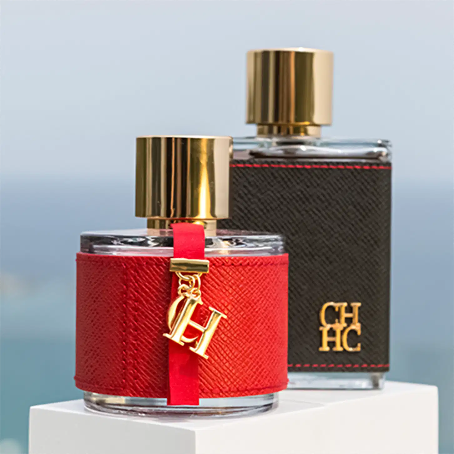 Dois frascos de perfume da marca Carolina Herrera, um vermelho e outro preto, com detalhes dourados e logotipo da marca. O frasco vermelho possui uma fita vermelha e um pingente dourado com as iniciais "CH".