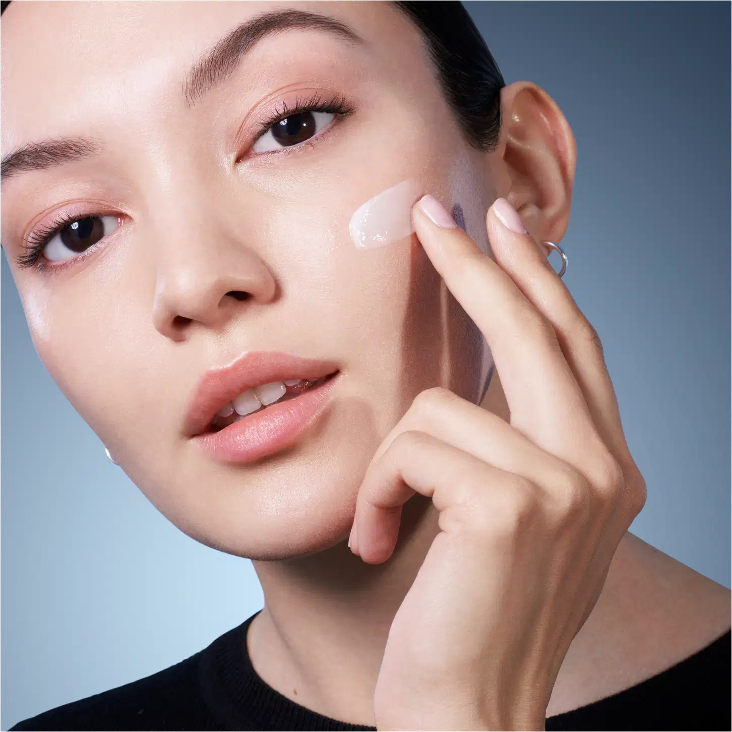 Retrato de uma mulher aplicando um produto de cuidados com a pele da marca Lancôme. Fundo azul claro, iluminação suave, expressão serena e concentrada.