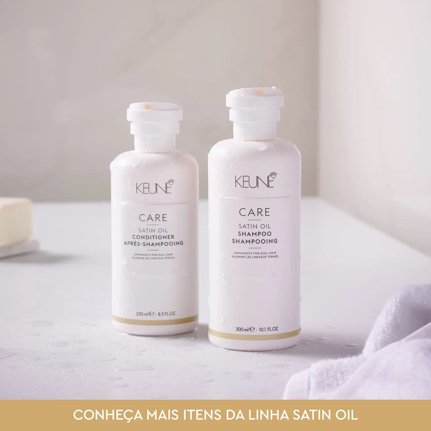 Dois frascos brancos da linha Satin Oil da Keune, contendo condicionador e shampoo. Os frascos possuem rótulos com o nome da marca e do produto, em cores branca e dourada.