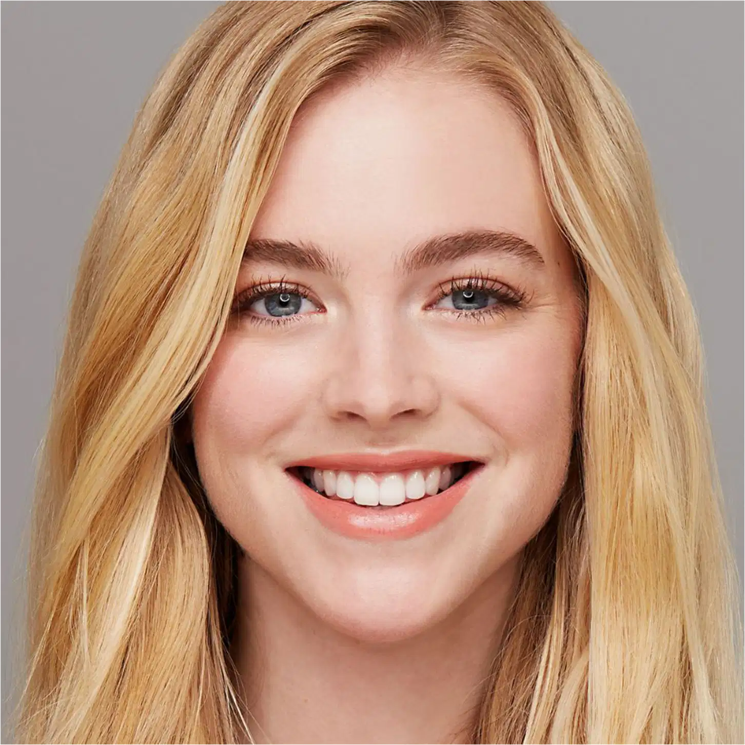 Close-up do rosto sorridente de uma mulher loira jovem com olhos azuis e dentes brancos. Ela tem uma expressão alegre e um visual natural e atraente.