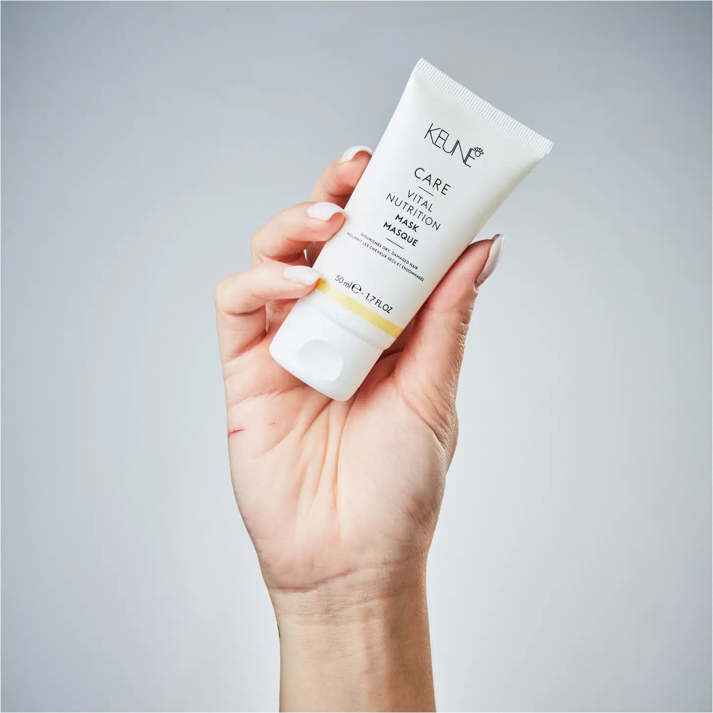 Mão segurando um tubo de máscara facial da marca Keune, em um ambiente minimalista e monocromático, sugerindo um contexto de cuidados com a pele e beleza.