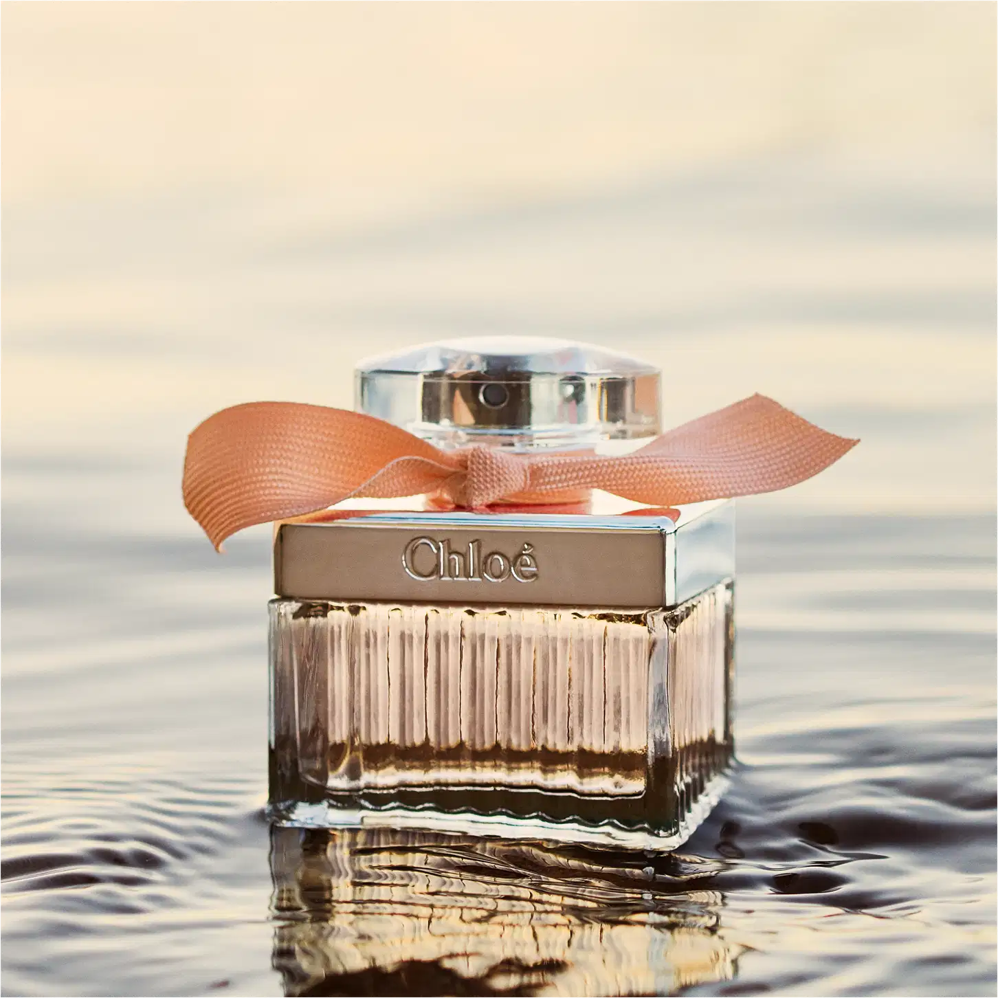 Frasco de perfume Chloé com tampa prateada e laço laranja, em embalagem de vidro com listras verticais, exibindo o nome da marca em destaque.