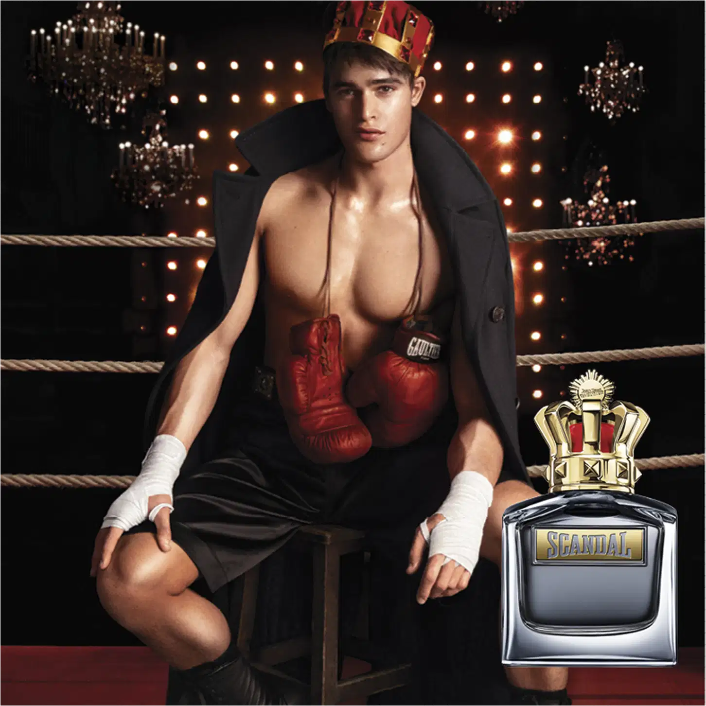 O ambiente retratado é um ringue de boxe iluminado por luzes brilhantes, com um homem musculoso vestindo luvas vermelhas e um casaco preto, segurando um frasco de perfume da marca Jean Paul Gaultier, sugerindo uma associação entre o produto e o esporte.