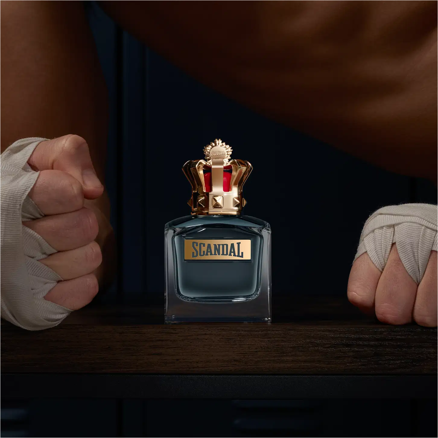 Frasco de perfume Jean Paul Gaultier Scandal, com tampa dourada em formato de coroa e rótulo em fundo azul escuro. Embalagem elegante e sofisticada, com design marcante e distintivo da marca.