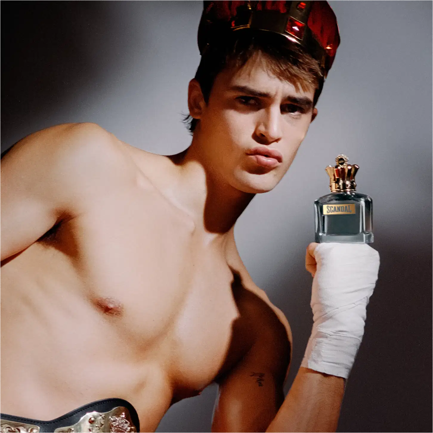 Ambiente escuro e dramático, com modelo masculino segurando um frasco de perfume da marca Jean Paul Gaultier, destacando-se com sua expressão séria e pose desafiadora.