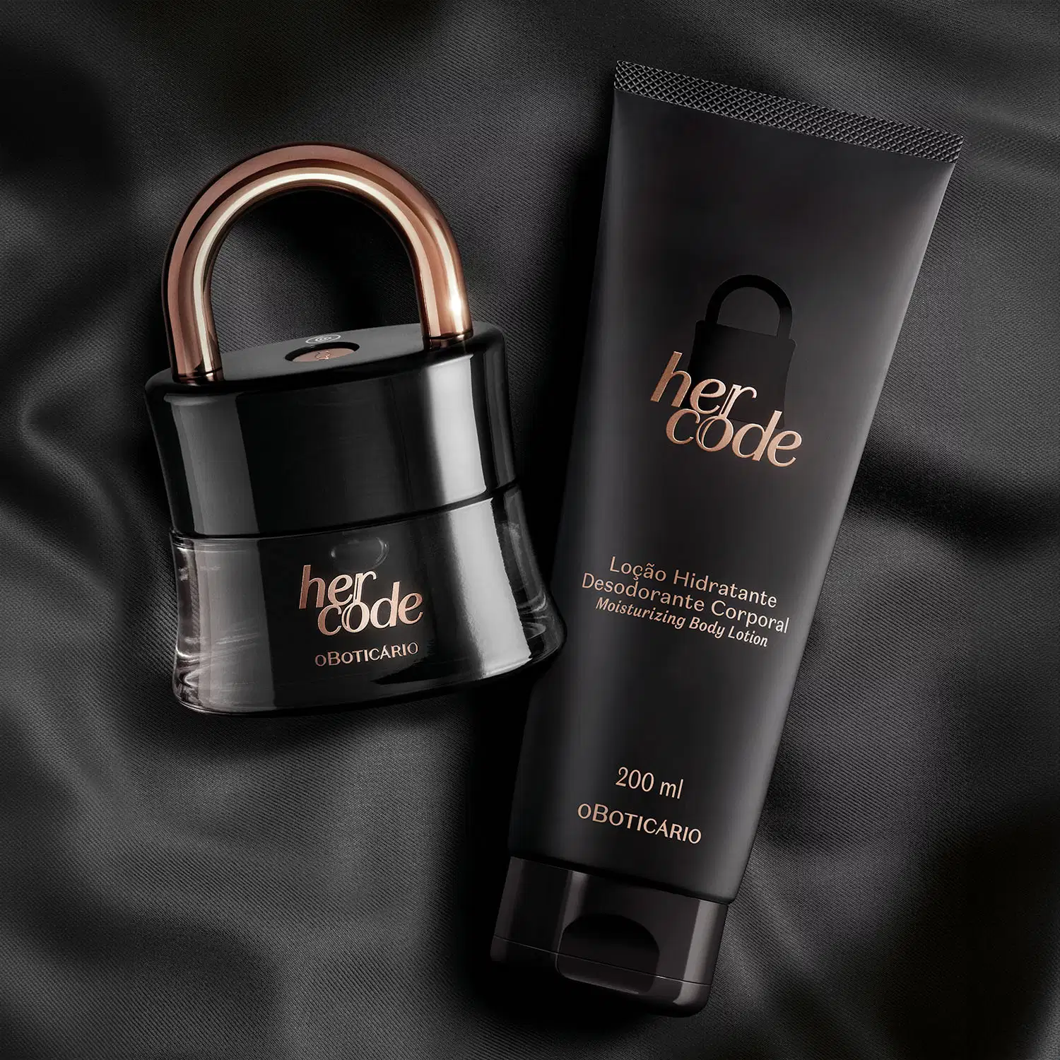 Dois itens da marca Her Code em embalagens pretas com letras douradas, sendo um vidro em formato de cadeado com arco dourado  contendo Eau de Parfum e uma bisnaga contendo loção hidratante corporal. O fundo da imagem é um tecido preto.
