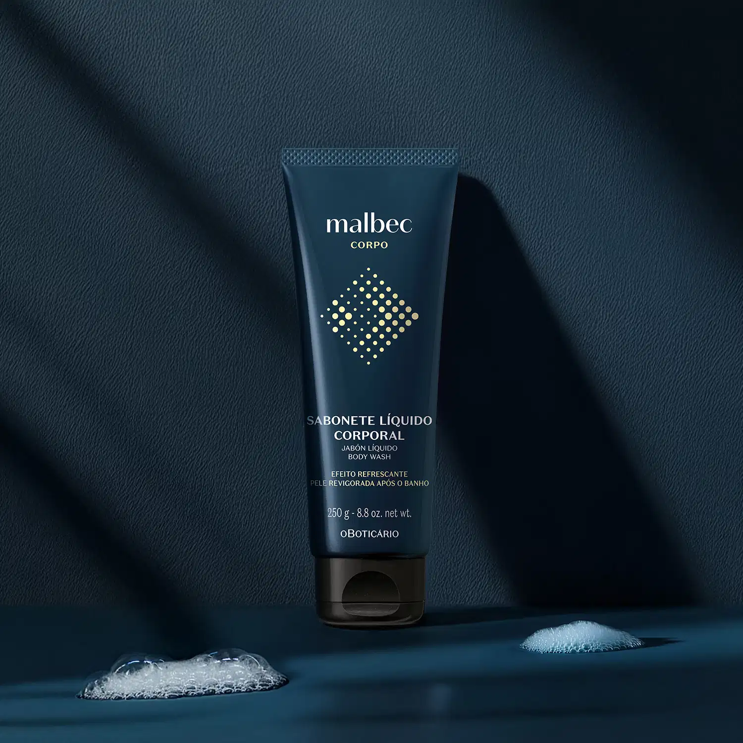 Imagem de um tubo azul-marinho de sabonete líquido corporal da marca Malbec sobre superfície escura com reflexos e sombras, em ambiente minimalista e monocromático.