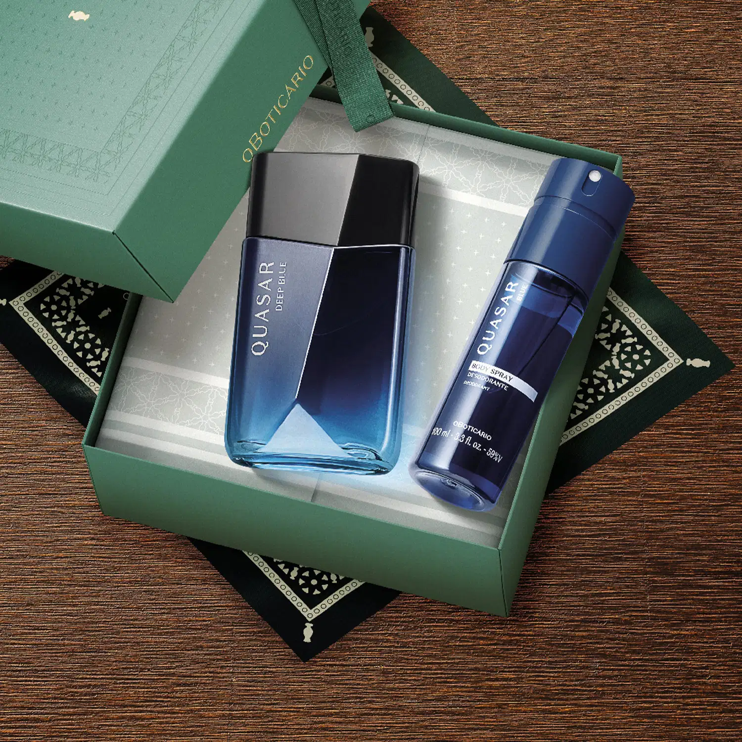 Imagem de uma caixa verde contendo um frasco de desodorante colônia em tons de azul e um body spray azul sobre uma superfície de madeira escura com detalhes decorativos pretos.