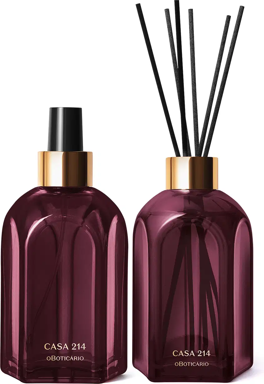 A imagem mostra dois vidros retangulares roxos, com rótulo em dourado. Um deles é o aromatizador de ambientes com válvula spray preta e o outro é um difusor com tampa dourada que acompanha varetas de madeira. Ambos são a linha Casa 214 Figo Magnífico.