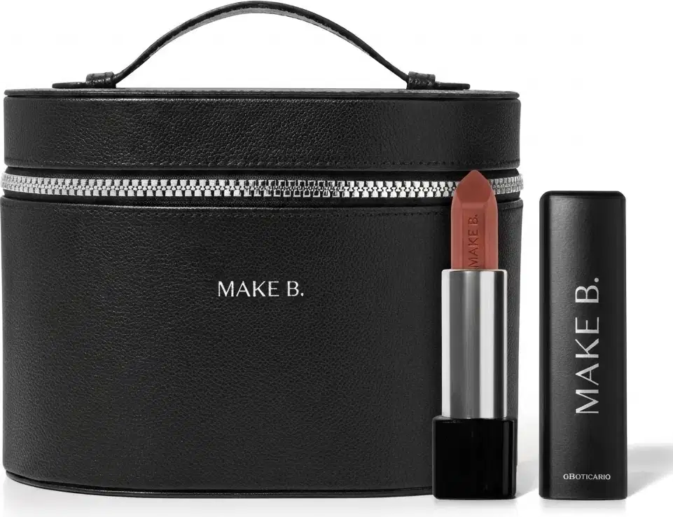Frasqueira de maquiagem preta com zíper e alça, com aspecto rígido e estruturado feita em tecido tipo couro e logo Make B. em hot stamping prata e um batom vermelho aberto com a bala visivel, corpo prateado e base preta com tampa preta ao lado.